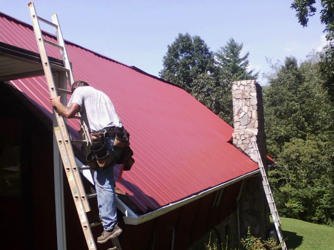 Roofer on a ladder beside a red metal roof and stone chimney — Soffit & Fascia Repair by Roofing Brothers in South Windsor